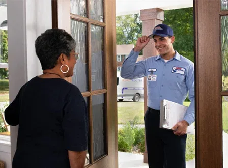 A uniformed roofer holding a clipboard stands at a residential doorway speaking with a homeowner.